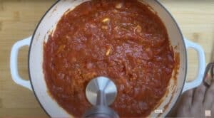 Blending homemade tomato sauce in a white pot with an immersion blender on a wooden table.